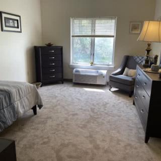 A cozy bedroom with light carpet, featuring a bed, dresser, and a chair by the window.