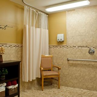 A well-lit bathroom with shower curtain, chair, and handrails on tiled walls.