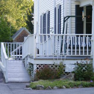 A white house with a front porch, featuring a ramp and a swing, surrounded by greenery.