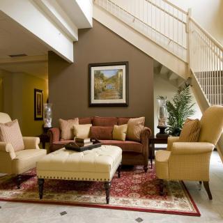 A cozy living room featuring a sofa, armchairs, and a coffee table beneath a staircase, decorated with artwork and a patterned rug.