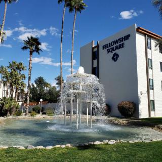 A fountain in front of an apartment building with palm trees and a clear blue sky.