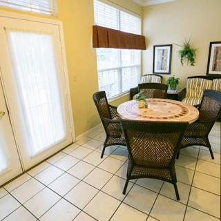 A cozy dining area with a round table and wicker chairs, illuminated by large windows.