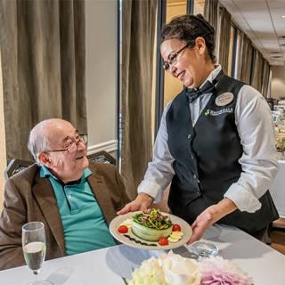A waitress serves a meal to an elderly man at a restaurant, as they share a friendly interaction.