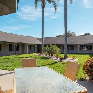A sunny courtyard with a patio table and chairs surrounded by palm trees and a well-maintained lawn.
