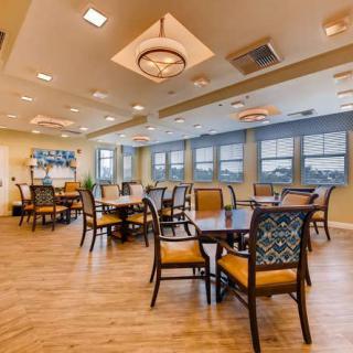 A spacious dining area with multiple tables and chairs, well-lit by ceiling lights and featuring large windows.