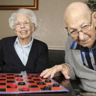 An elderly couple playing a game of checkers indoors, both smiling.