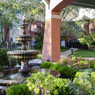 A serene garden featuring a decorative fountain surrounded by lush greenery and vibrant flowers, set against a backdrop of brick architecture.