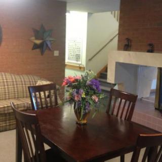 A cozy dining area with a wooden table and a vase of flowers, adjacent to a living room featuring a sofa and fireplace.