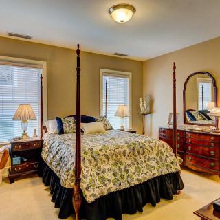 A cozy bedroom with a four-poster bed, dresser, and mirror, warmly lit by lamps and featuring two windows.