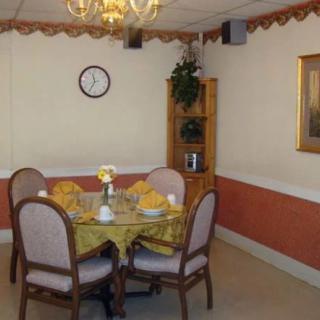A cozy dining room setup featuring a round table with four chairs, adorned with a tablecloth and flower centerpiece. A clock hangs on the wall above a wooden corner shelf with decorative items.