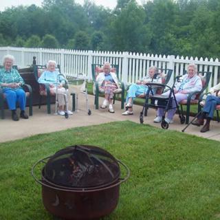 A group of people sitting outside on chairs near a fire pit, with a white fence and greenery in the background.