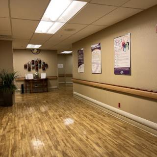 A view of a well-lit hospital hallway featuring wooden flooring, informational posters on the walls, and potted plants.