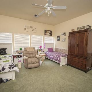 A cozy bedroom with two beds, an armchair, and a wardrobe, featuring a ceiling fan and ample natural light from the windows.