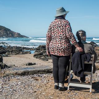 Two people enjoy a scenic view by the ocean, one seated in a chair and the other standing beside them.