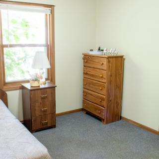 A cozy, minimalist bedroom featuring a wooden dresser and bedside table with a lamp.