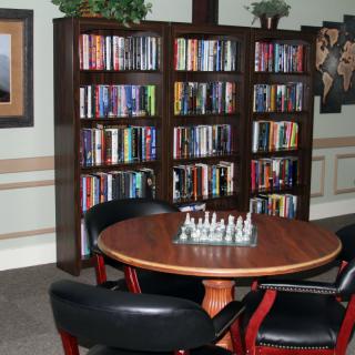A cozy library corner featuring a round table with a chessboard, surrounded by bookshelves and chairs.