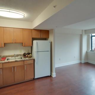 A compact apartment kitchen with wooden cabinets and hardwood flooring, featuring a refrigerator and window.