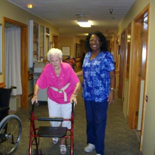 An elderly woman using a walker is accompanied by a nurse in a hallway of a care facility.