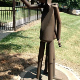 A metal abstract sculpture depicting a figure with one arm raised, set in an outdoor park.