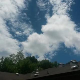 A view of fluffy clouds in a clear blue sky over a rooftop with trees in the background.