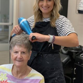 A hairstylist blow-drying a customer's hair while both are smiling in a salon.
