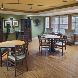 A cozy dining area with wooden floors, tables, and chairs complemented by natural light from large windows.