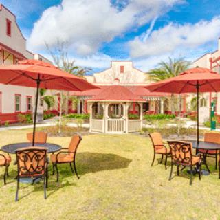 A sunny courtyard with tables, chairs, and umbrellas surrounded by buildings.