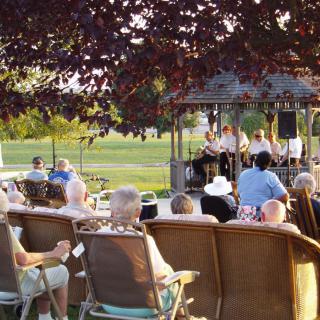 People enjoying an outdoor concert in a park, seated around a gazebo on a sunny day.