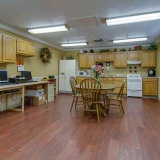 A multi-purpose room featuring a kitchen and office area with computers, a central wooden table, and chairs.