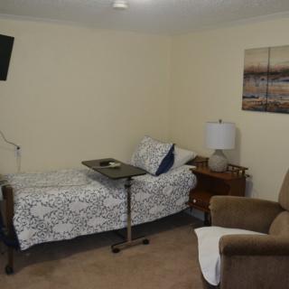 A cozy bedroom with a single bed, side table, armchair, and a television on the wall.