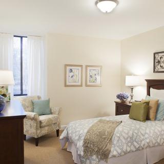 A cozy bedroom with elegant decor, featuring a neatly made bed, artwork on the walls, and a comfortable armchair.