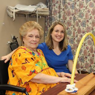 An elderly woman in a wheelchair, accompanied by a caregiver, smiling in a nursing home setting.