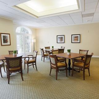 A cozy dining room with several tables and chairs arranged on a carpeted floor, illuminated by natural light through large windows.