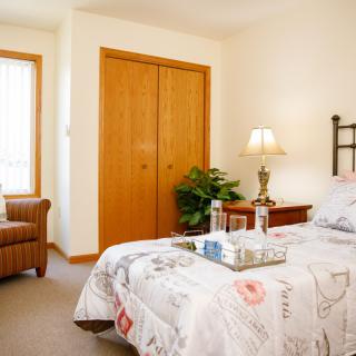 A cozy bedroom with a neatly made bed, bedside lamp, and comfortable chair near a window.