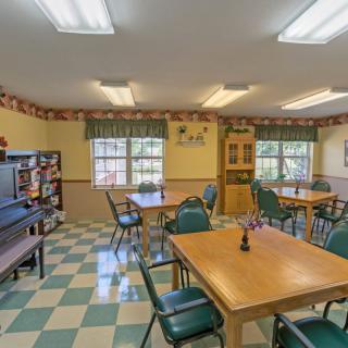 A cozy dining room with tables and chairs, featuring a bookshelf and large windows.