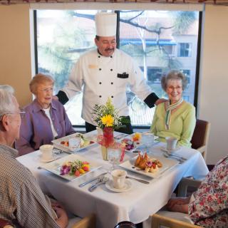 A chef interacts with elderly guests enjoying a meal at a restaurant.