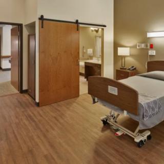 A hospital room featuring a bed and medical equipment, with warm lighting and wooden flooring.