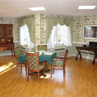 A cozy dining area with a round table and four chairs, set on a wooden floor with large windows and floral wallpaper.