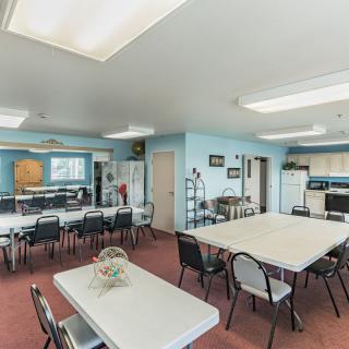 A community room featuring tables, chairs, and a small kitchen area with bright lighting.