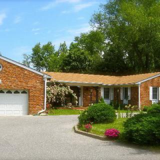 A brick house with a surrounding garden and trees, featuring a driveway leading to an attached garage.
