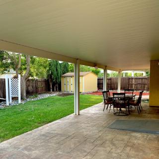 A spacious outdoor patio with a dining table under a covered area, overlooking a lush green lawn and wooden fence.