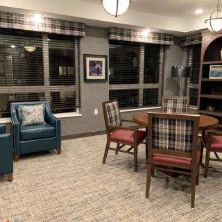 A cozy living room with a round table, armchair, and plaid-upholstered chairs under warm indoor lighting.