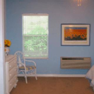 A cozy bedroom with blue walls, featuring a window with blinds, a dresser, a chair, and a framed painting on the wall.