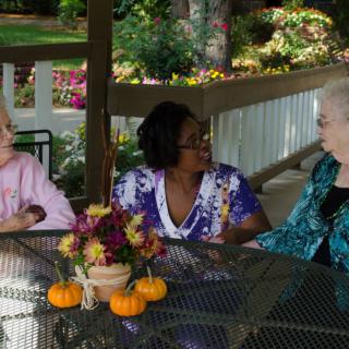 Three women sitting at an outdoor table having a conversation, surrounded by small pumpkins and flowers.