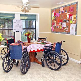 A cozy community area in a nursing home with wheelchairs around a decorated table and a colorful bulletin board on the wall.