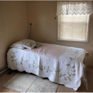 A minimalist bedroom with a single bed covered in a quilt, softly lit by natural light from a window.