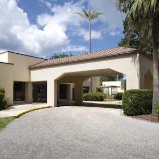 A modern architectural building featuring a covered entrance, surrounded by lush greenery and palm trees under a partly cloudy sky.