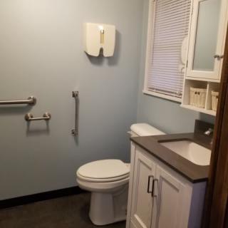 A clean and accessible bathroom with a sink, mirror, toilet, handrails, and a window.