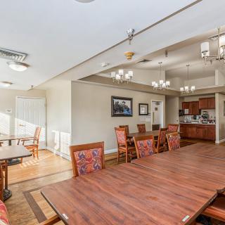 A spacious dining area with a large wooden table, chairs, and adjacent kitchen, well-lit with natural light and chandeliers.