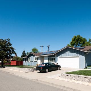A suburban house with solar panels on the roof and a car parked in the driveway under a clear blue sky.
